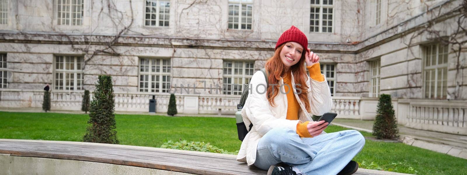 Young smiling redhead girl sits on bench and uses smartphone app, reads news online, watches video on mobile phone while relaxed in park by Benzoix