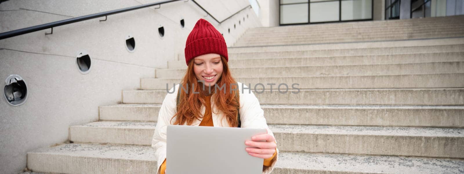 Smiling redhead girl, student sits on stairs outdoors and uses laptop, connects to public wifi in city and works on project, uses internet on computer.