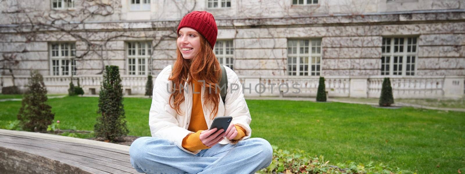 Young smiling redhead girl sits on bench and uses smartphone app, reads news online, watches video on mobile phone while relaxed in park.