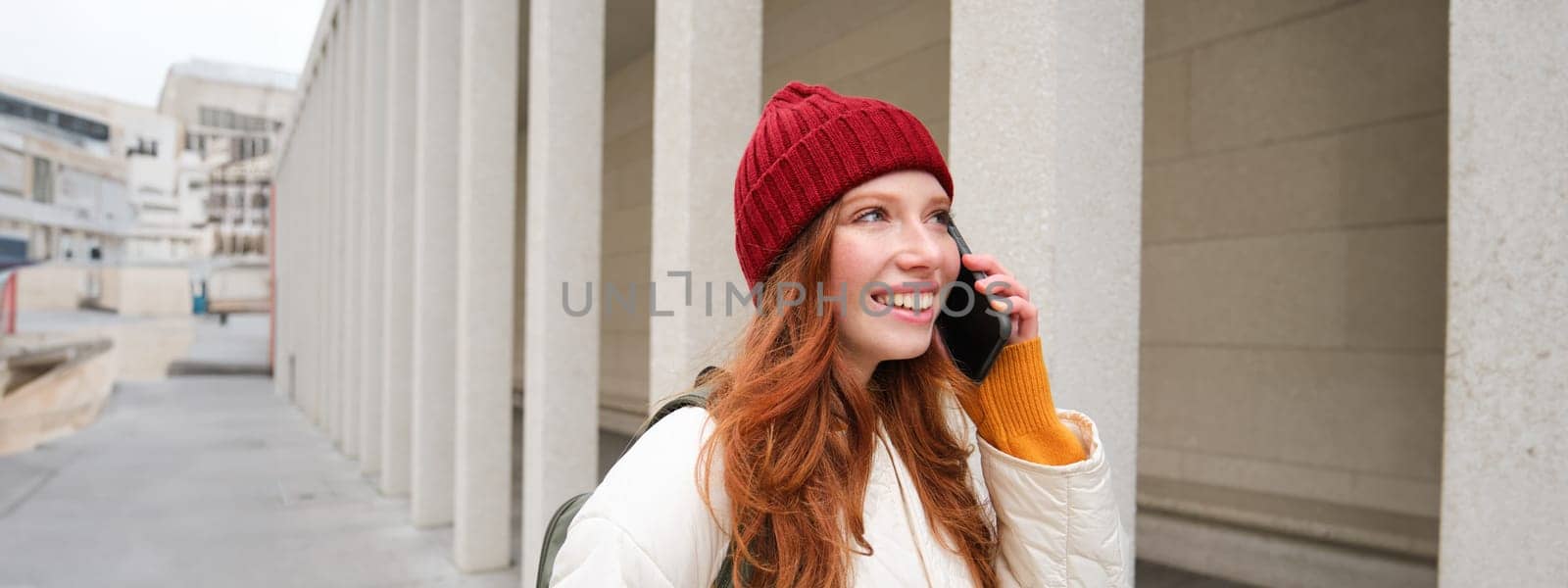 Mobile broadband and people. Smiling young redhead woman walks in town and talks on mobile phone, calling friend on smartphone, using internet to make a call abroad by Benzoix