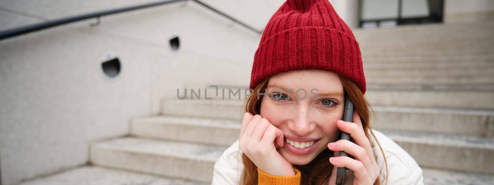 Young stylish redhead girl in red hat, sits on street and talks on mobile phone, has telephone conversation, rings her friend while relaxes outdoors by Benzoix