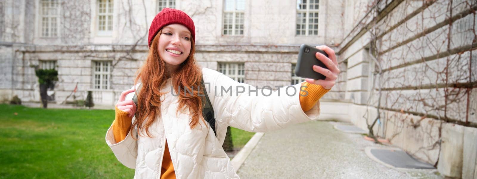 Portrait of happy girl tourist, takes selfie on smartphone in front of historical building, posing for photo on mobile phone camera.