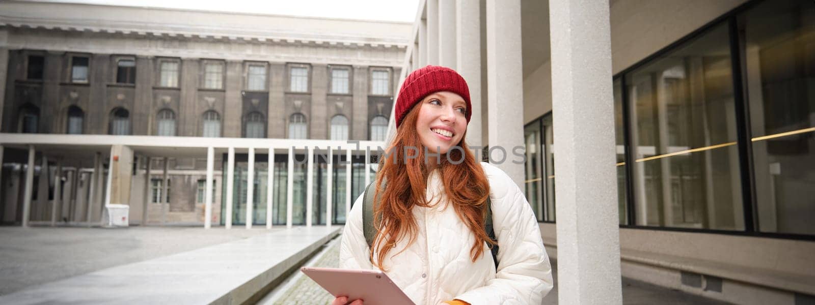 Stylish ginger girl, tourist walks with digital tablet around city, woman connects to iternet on her gadget, looking up information, texting message by Benzoix