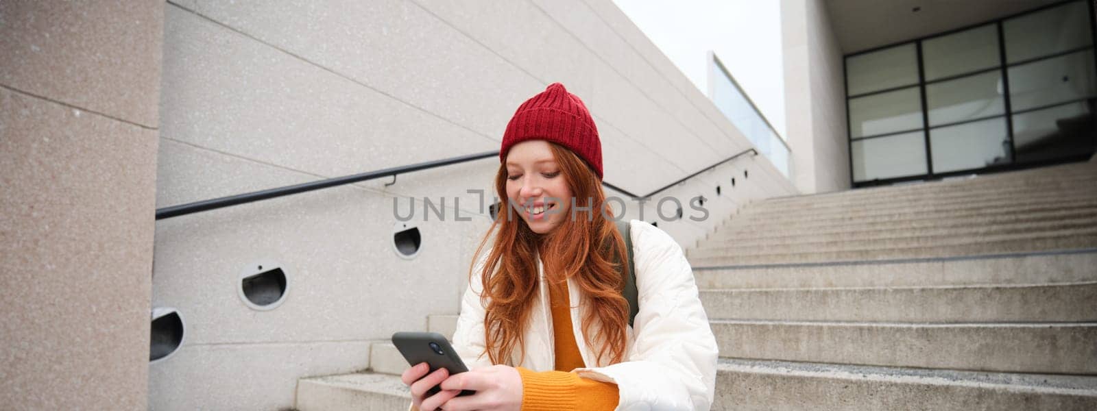 Redhead girl in red hat, sits on stairs and uses mobile phone. Modern woman holding smartphone, texting message, using telephone application outdoors by Benzoix