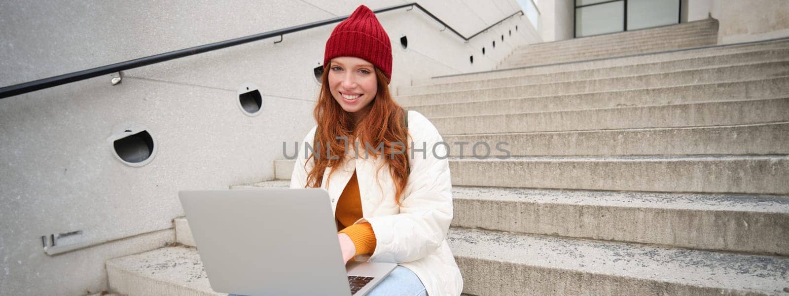 Smiling redhead girl, young woman typing on laptop keyboard, sitting outdoors on stairs with computer, working remote, doing her homework on fresh air by Benzoix