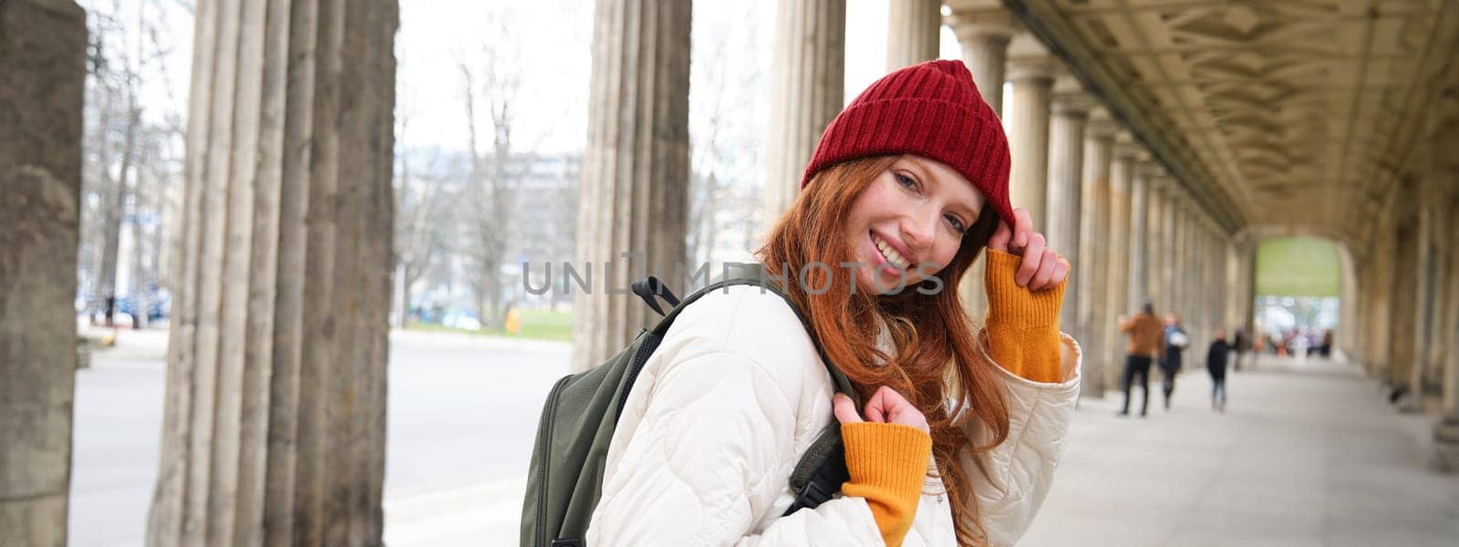Cute smiling girl walks around city in red hat and gloves, has backpack, tourist explores strees of european city by Benzoix