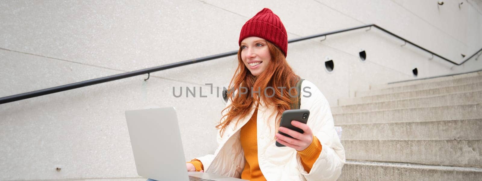 Smiling redhead woman with mobile phone and laptop, sitting on stairs outside building, connects to public wifi, using smartphone and computer.