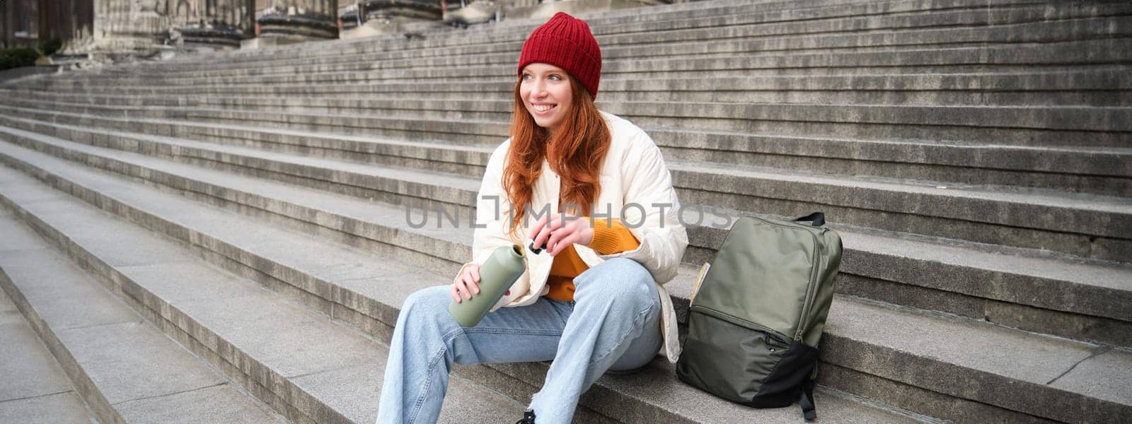 Smiling tourist, girl sits on stairs, rests on staircase, takes thermos from backpack, drinks hot coffee from flask.