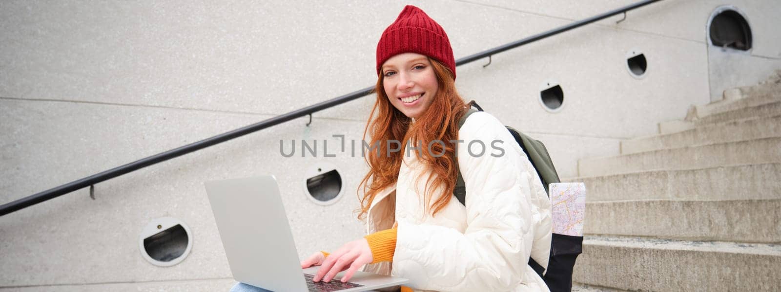 Portrait of young girl traveller, sitting with backpack and map of city, working on laptop, connecting to public wifi and sitting on stairs outdoors, using internet to book hotel room.