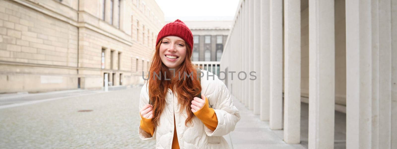 Female tourist in red hat with backpack, sightseeing, explores historical landmarks on her trip around europe, smiling and posing on street.