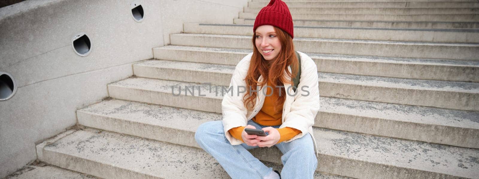Hipster ginger girl, redhead woman sits on stairs with smartphone, waits for someone and messages on social media on mobile phone app. People and technology concept