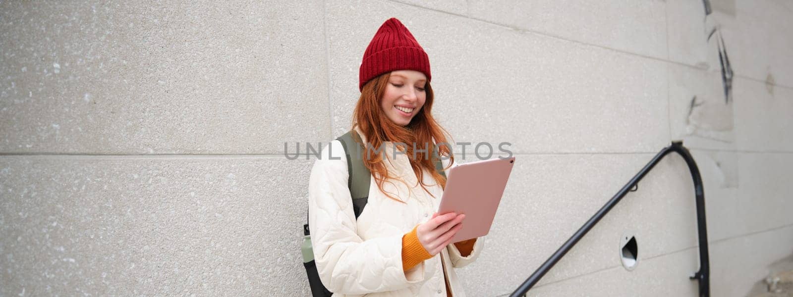 Young redhead woman with red hat, uses her digital tablet outdoors, stands on street with gadget, connects to wifi internet and searches for a location in internet by Benzoix