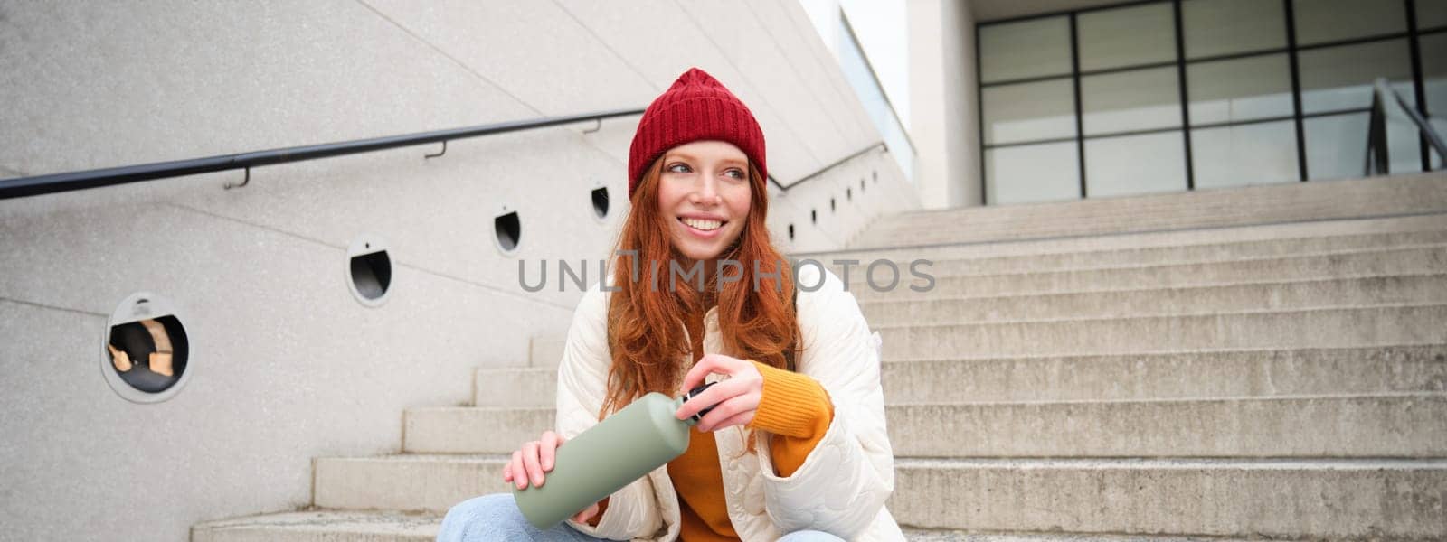 Smiling traveler, redhead girl tourist sits on stairs with flask, drinks hot coffee from thermos while travelling and sightseeing around foreign city, sits on stairs and rests by Benzoix