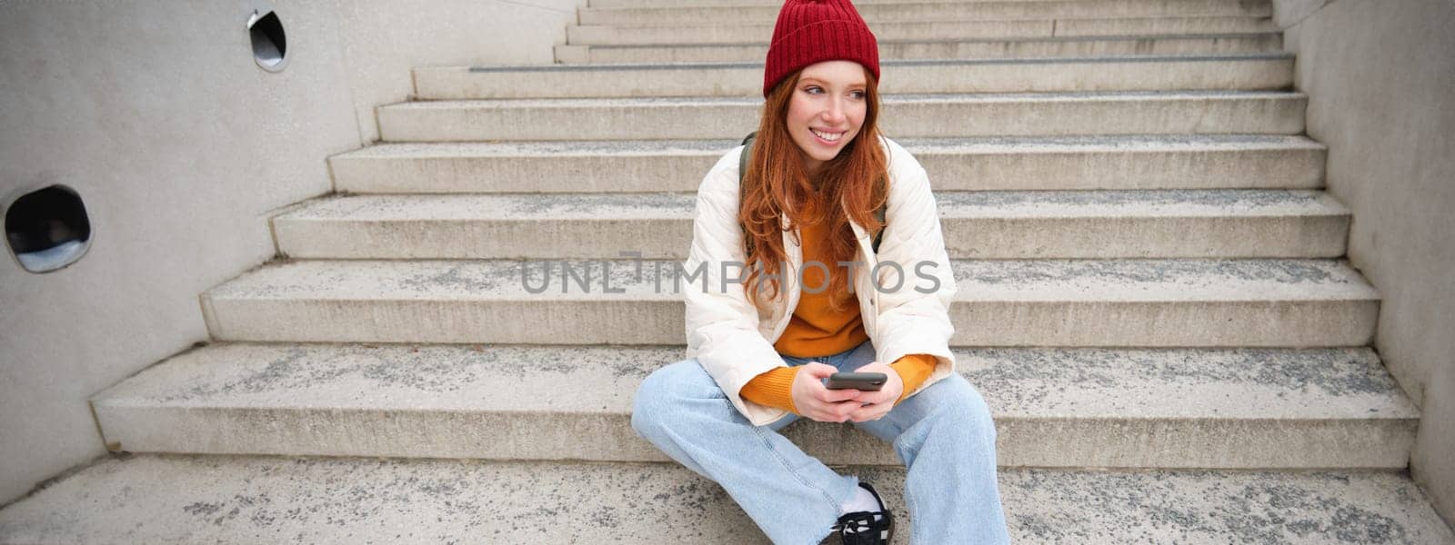 Hipster ginger girl, redhead woman sits on stairs with smartphone, waits for someone and messages on social media on mobile phone app by Benzoix