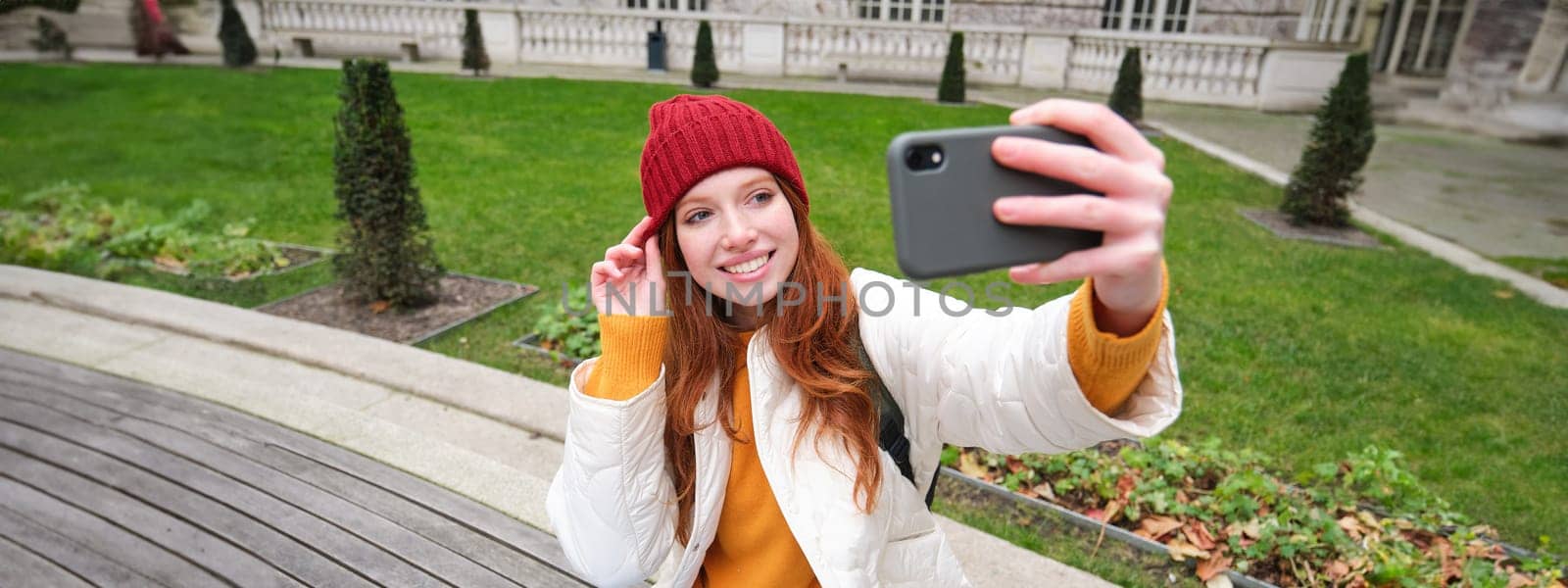 Young teen redhead girl sits on bench in park and takes selfie, makes a photo of herself with smartphone app, records vlog by Benzoix