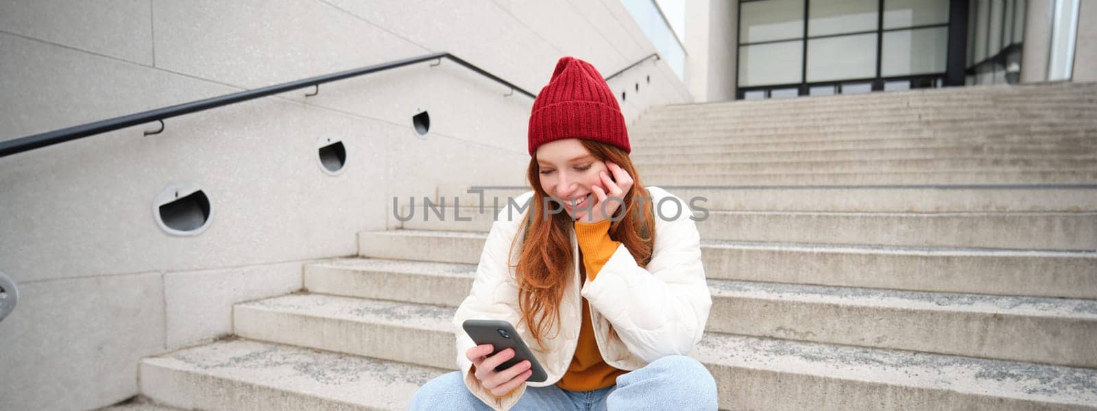 Beautiful redhead teen girl, sits on stairs and smiles, holds mobile phone, uses smartphone application, chats on telephone app by Benzoix