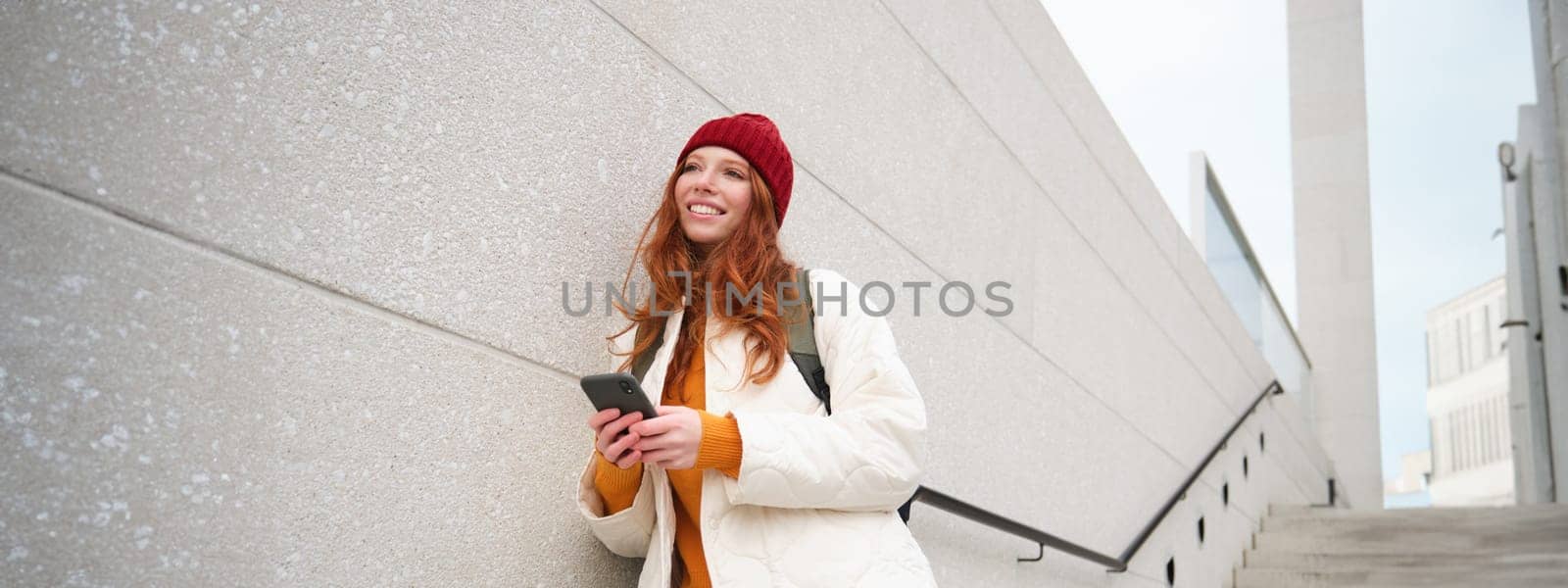 Happy girl student in red hat, holds smartphone, tourist looks at map app on her phone, explores sightseeing, texts message, looks for couchsurfing, rents place to stay online.
