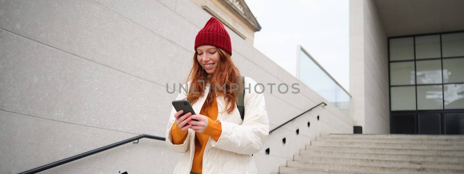 Redhead girl, young woman tourist with backpack, holds smartphone, looks for route on mobile application, searches for hotel on phone map, smiles happily by Benzoix