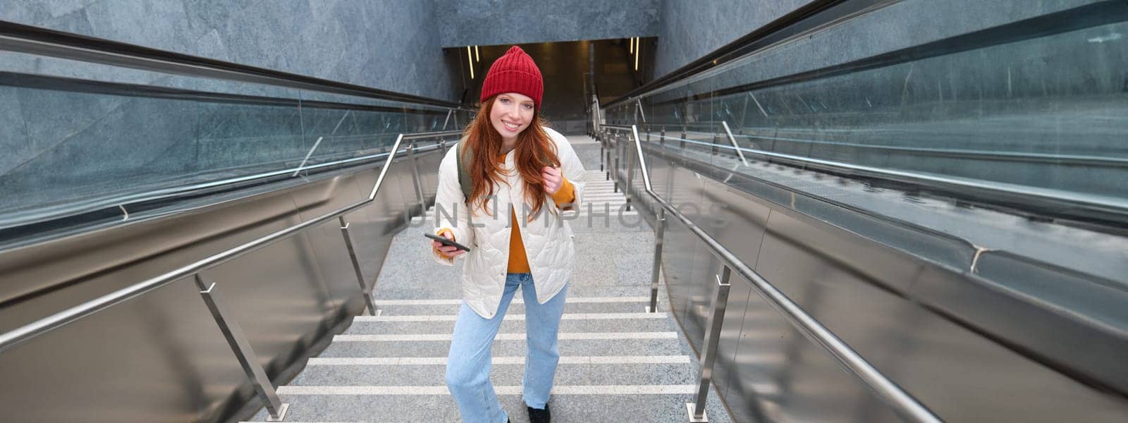 Portrait redhead girl tourist, goes up stairs with smartphone, follows route on mobile phone app, holds backpack and smiles.