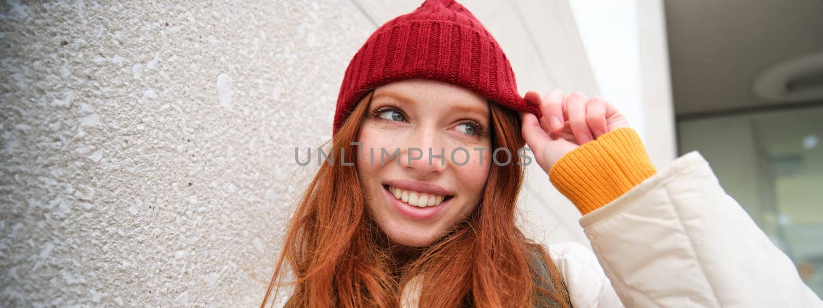 Stylish redhead girl in red hat, smiles and looks happy, poses outdoors on street, looks relaxed and lively.