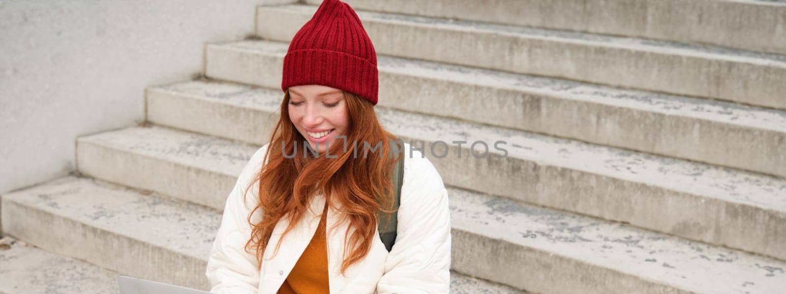 Smiling redhead girl, young woman typing on laptop keyboard, sitting outdoors on stairs with computer, working remote, doing her homework on fresh air.