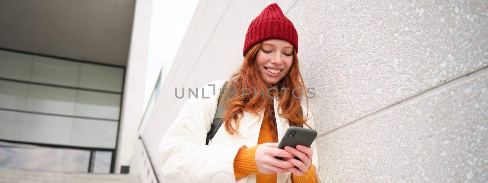 Happy girl student in red hat, holds smartphone, tourist looks at map app on her phone, explores sightseeing, texts message, looks for couchsurfing, rents place to stay online by Benzoix