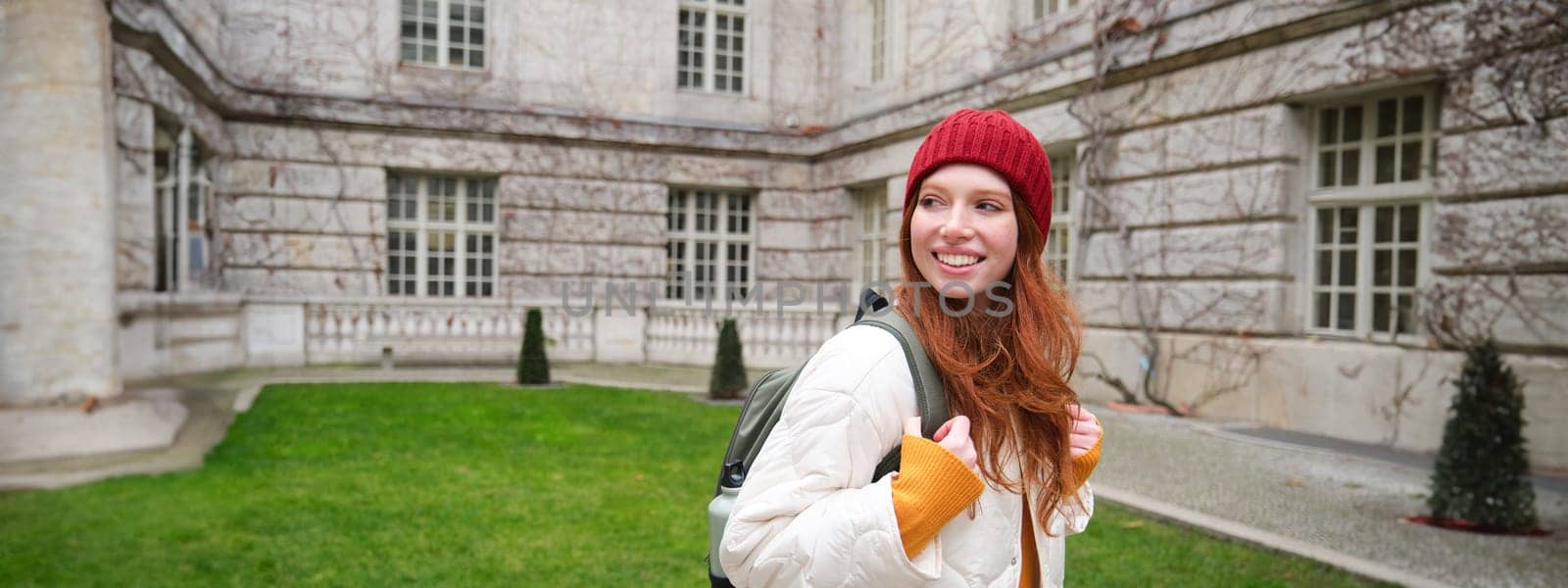 Beautiful redhead woman with backpack, tourist walking around city park and looks around, wears red hat and warm coat by Benzoix