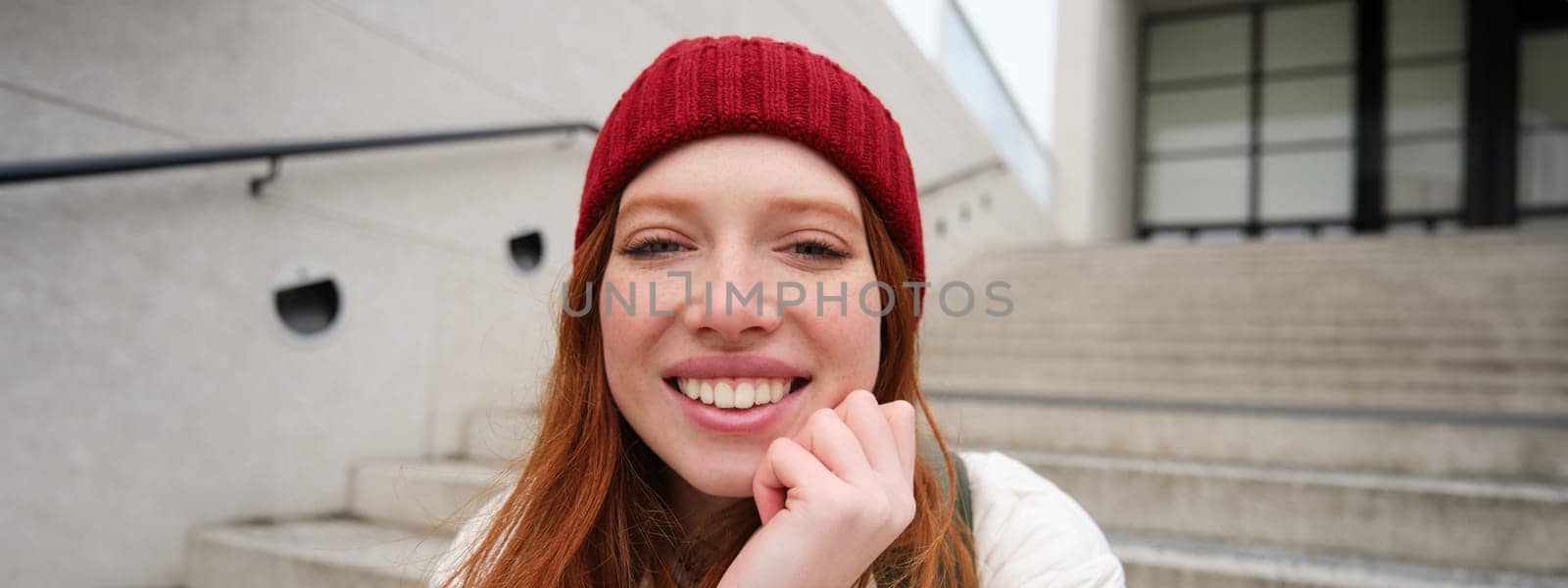 Beautiful redhead student, girl in red hat, smiles sincere, looks happy and relaxed, sits on stairs outdoors. Copy space