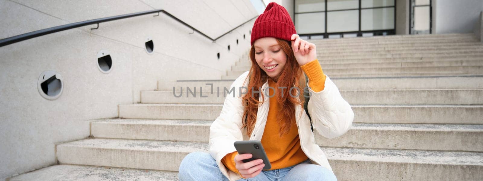 Stylish european girl with red hair, sits on public stairs with smartphone, places online order, sends message on mobile phone social app, smiles happily by Benzoix