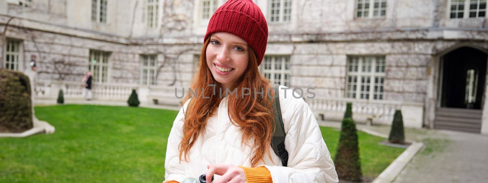 Portrait of happy girl traveler, drinking hot tea from thermos while walking and exploring city. Tourist with flask smiling at camera by Benzoix