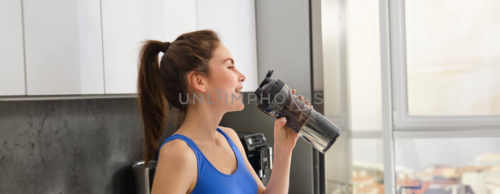 Image of fit and healthy, fitness woman in sportswear, drinking water, holding bottle shaker and standing in kitchen, staying hydrated after workout.