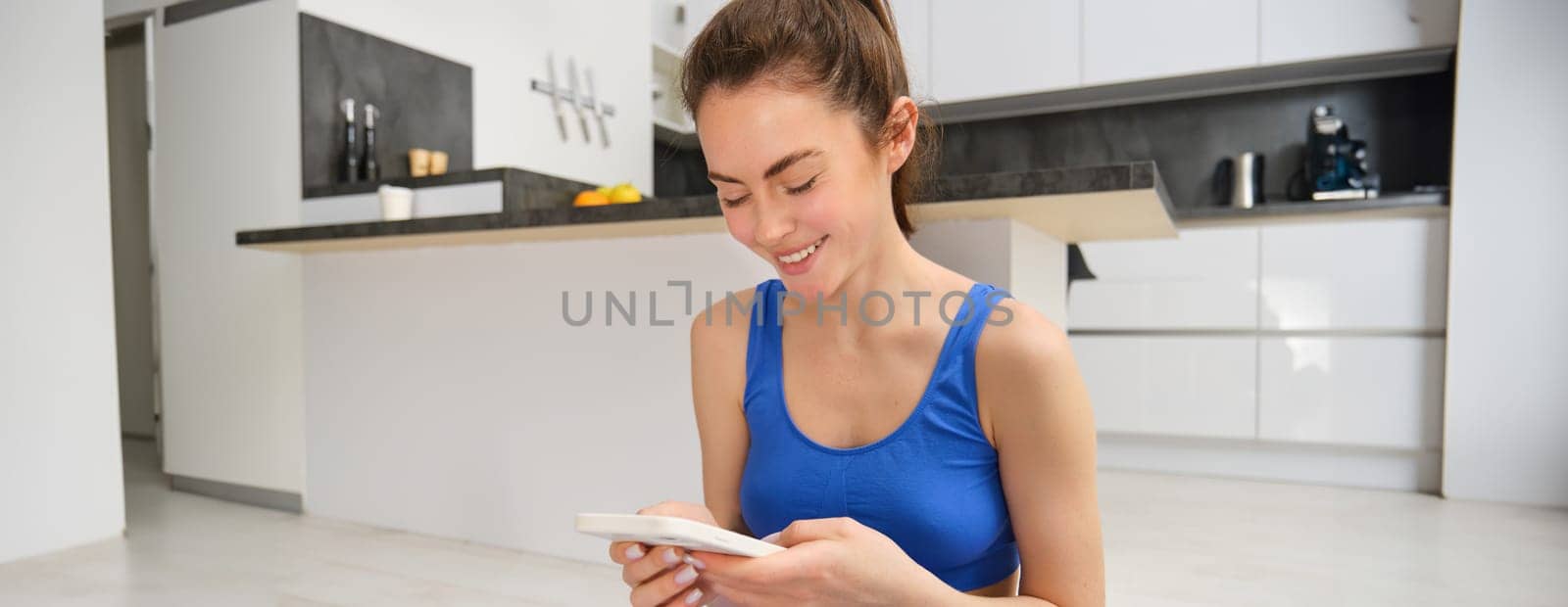 Close up portrait of sporty girl in sportsbra, holds smartphone, workout from home, looking at exercises on mobile phone. Wellbeing concept