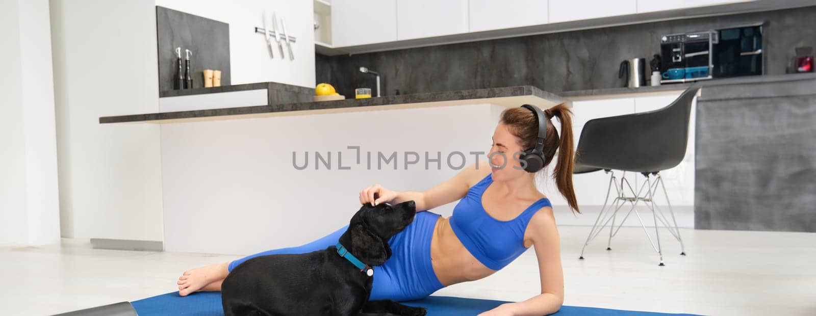 Portrait of happy young woman, tried to workout at home, laying on yoga mat in activewear, playing with her dog, listening music in wireless headphones by Benzoix