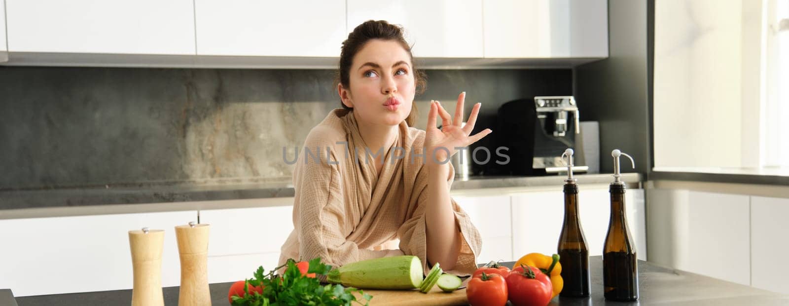 Portrait of beautiful young woman in bathrobe, cooking meal for family, shows chefs kiss, okay sign, making food, preparing vegetarian dinner, chopping vegetables.