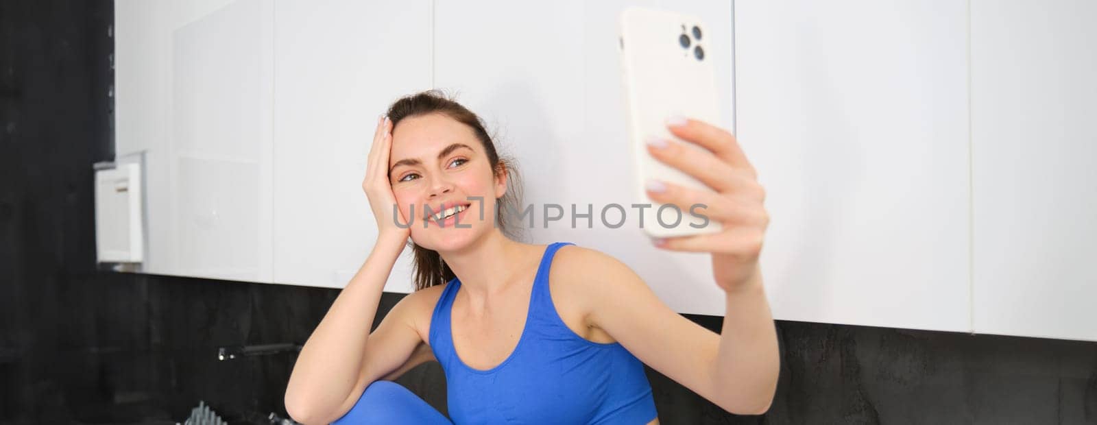 Image of fit and healthy, smiling fitness girl, posing for selfie on mobile phone, holding smartphone, looking at screen, taking photos in sportsbra and leggings.