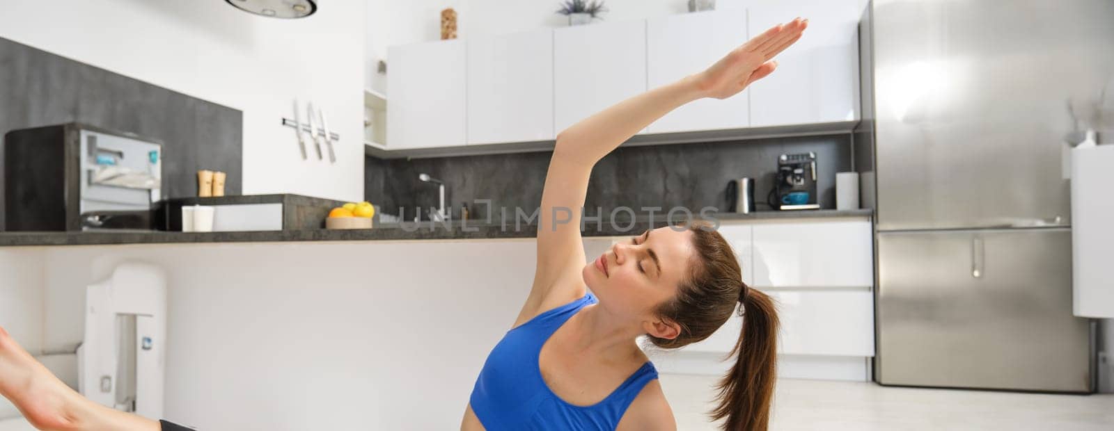 Image of young concentrated woman, doing sports, workout from home in blue sportsbra and legging, sits on yoga mat and stretches, practice relaxing asanas by Benzoix