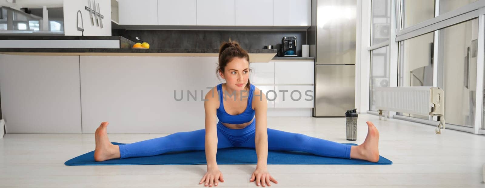 Fitness and workout concept. Young woman stretching her legs, doing exercises at home on yoga mat, doing splits on floor in living room.