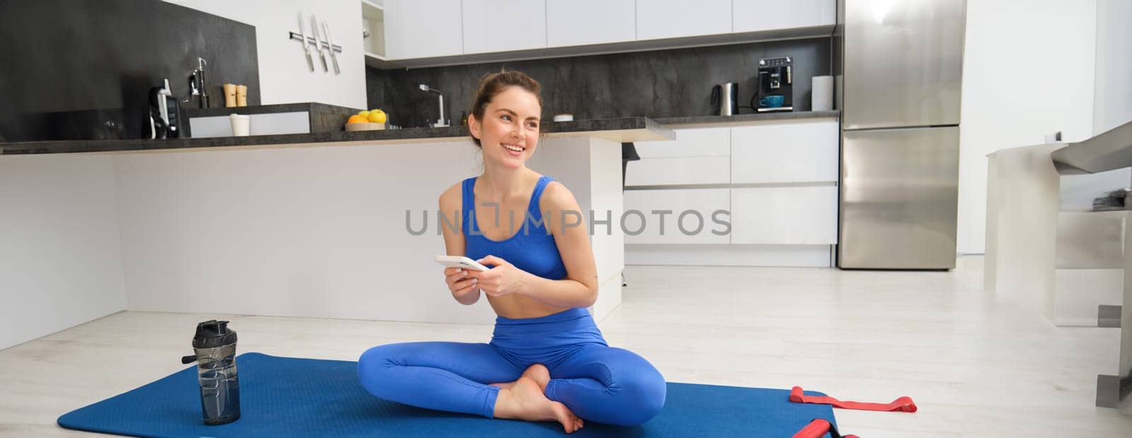 Sport and wellbeing. Young sportswoman does exercises at home, sits in yoga pose, holds smartphone, follows online training videos during her workout indoors.