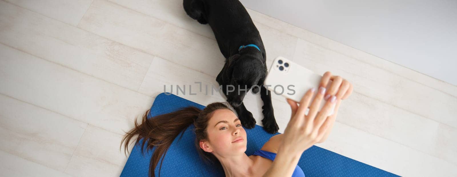 Portrait of beautiful sportswoman, fitness girl lying on floor at home with her dog, taking selfie with puppy, wearing workout clothes.
