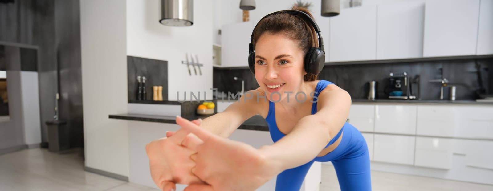 Portrait of beautiful, fit woman in blue leggings and sportsbra, stretching her arms, doing fitness workout, aerobics exercises at home, listening music in wireless headphones by Benzoix