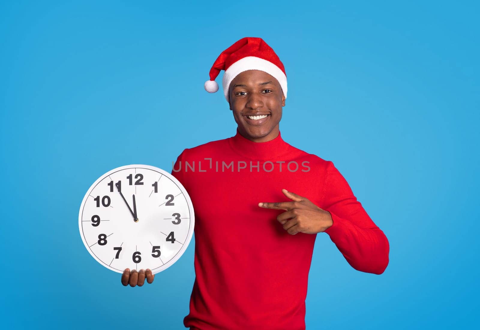 Black Man In Festive Santa Hat Pointing Finger At Clock Standing Against Blue Backdrop In Studio, Counting Down Minutes To Upcoming Year. Excitement Of Christmas And New Year Festivities
