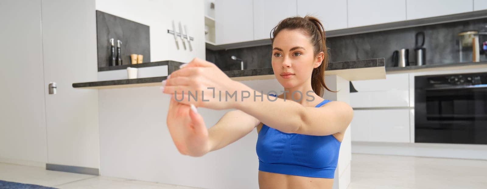 Young woman does workout from home, sits on yoga mat and stretches her hands, fitness training session in living room by Benzoix