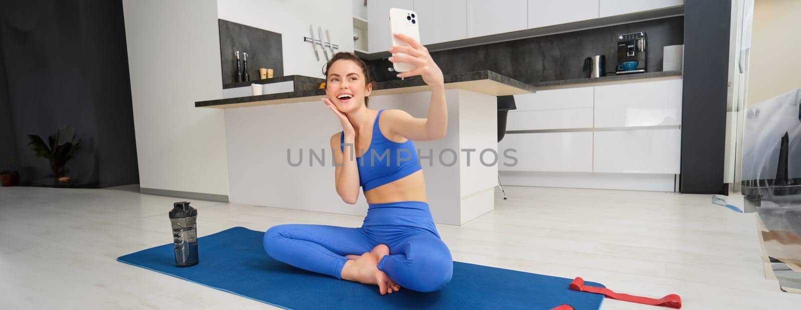 Sport and lifestyle concept. Young woman doing workout at home on yoga mat, makes fitness vlog with smartphone, takes selfie on mobile phone by Benzoix