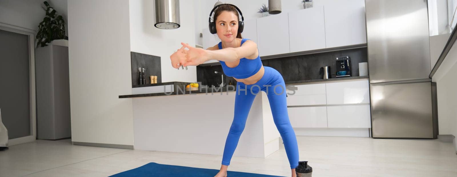 Portrait of beautiful, fit woman in blue leggings and sportsbra, stretching her arms, doing fitness workout, aerobics exercises at home, listening music in wireless headphones by Benzoix