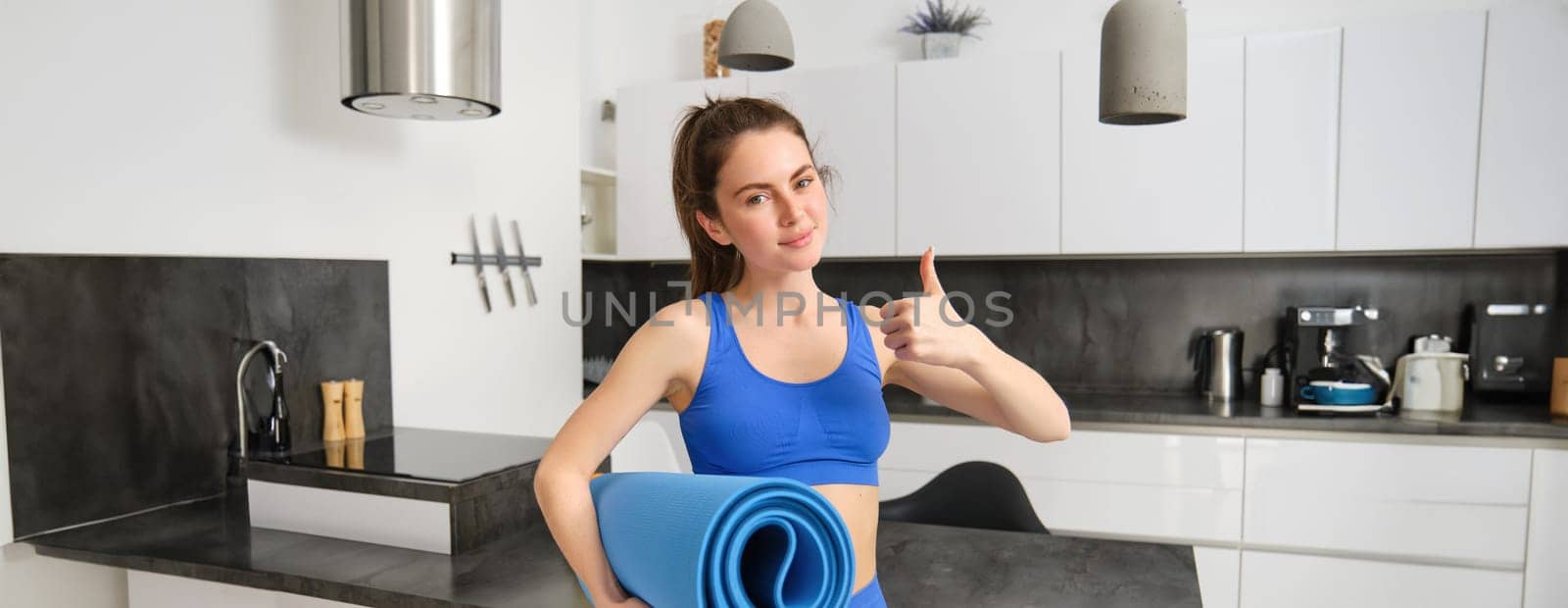 Attractive, sporty woman in blue activewear, shows thumb up, holds rubber yoga mat, workout at home, doing sport exercises in living room.