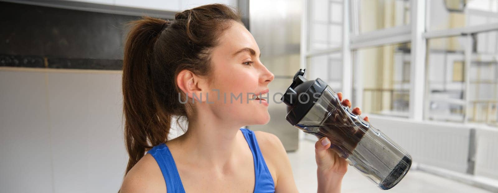 Beautiful young sportswoman drinks water, stays hydrated during workout training at home, doing fitness exercises in living room by Benzoix