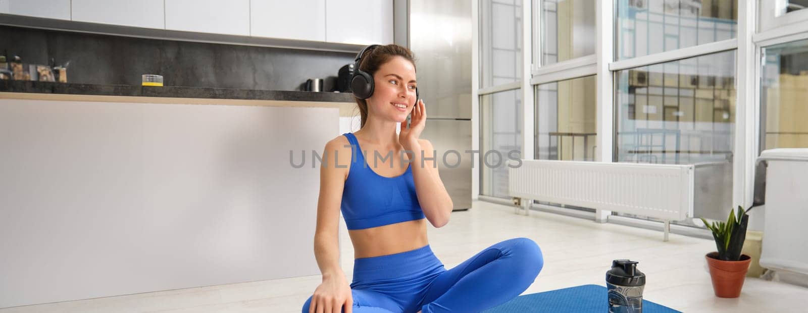 Young sporty athletic fitness trainer instructor woman wear blue tracksuit spend time in home gym use mobile phone headphones listen music. Workout sport concept by Benzoix