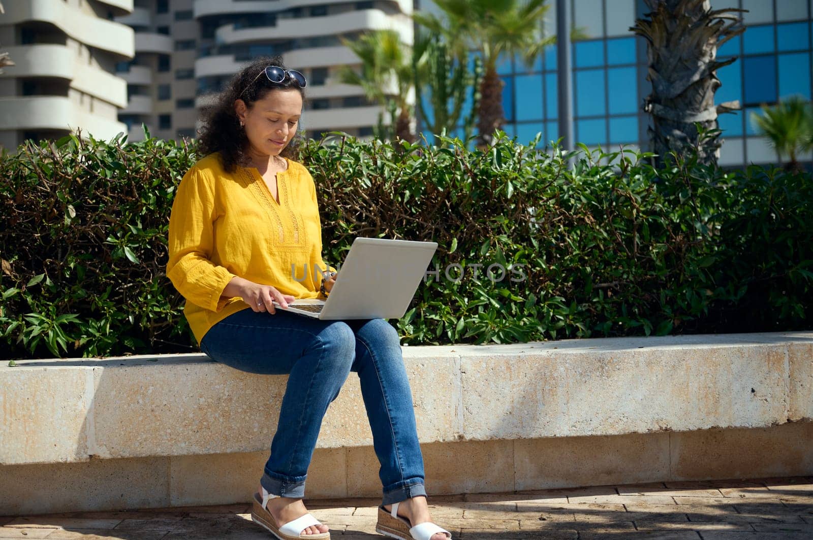 Full length portrait of multi-ethnic confident multitasking female freelancer, entrepreneur using laptop, working remotely, planning start-up or new business project, sitting on a stone bench outdoor.