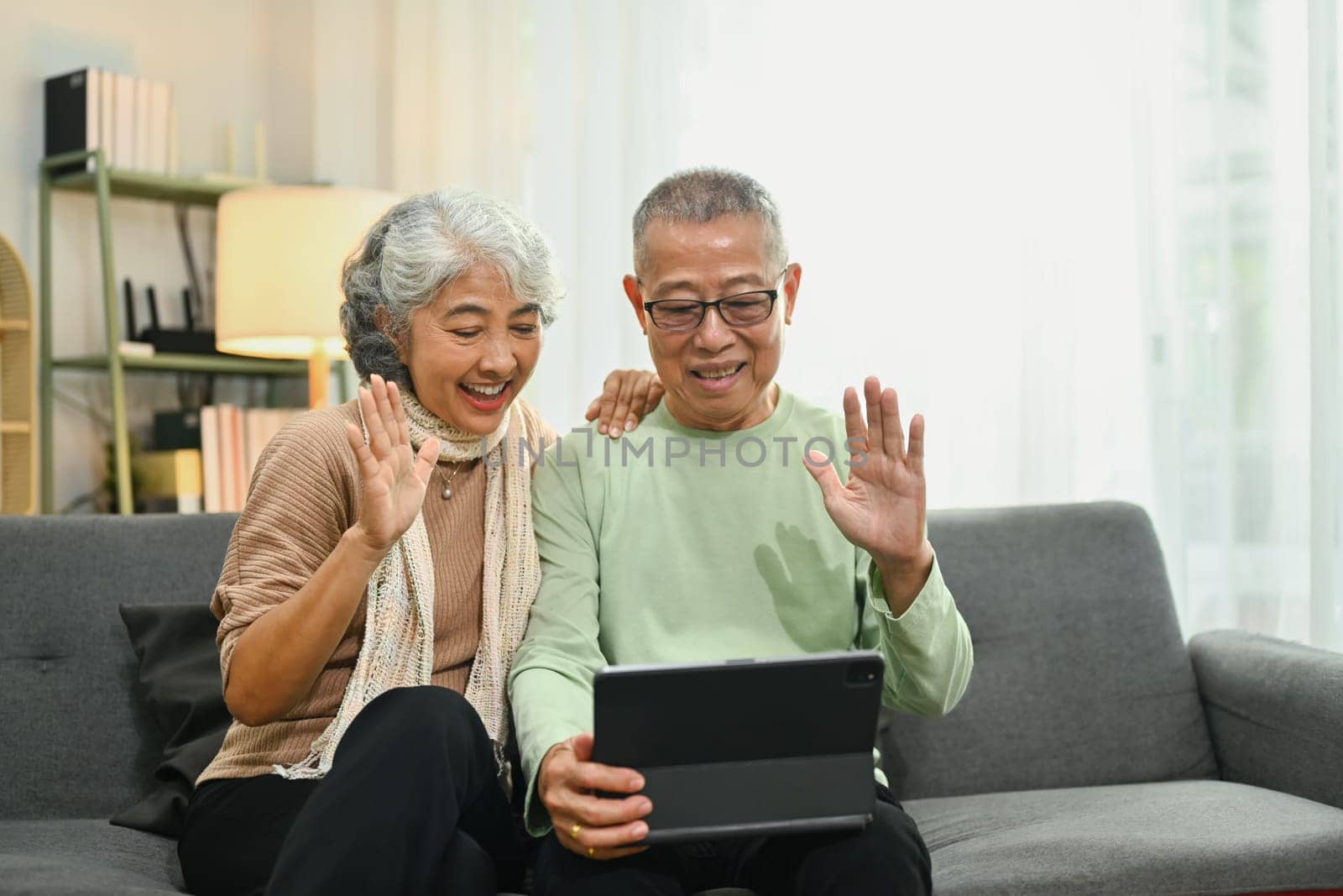 Cheerful elderly man waving hand speaking on video call talking to family while resting on armchair at home.