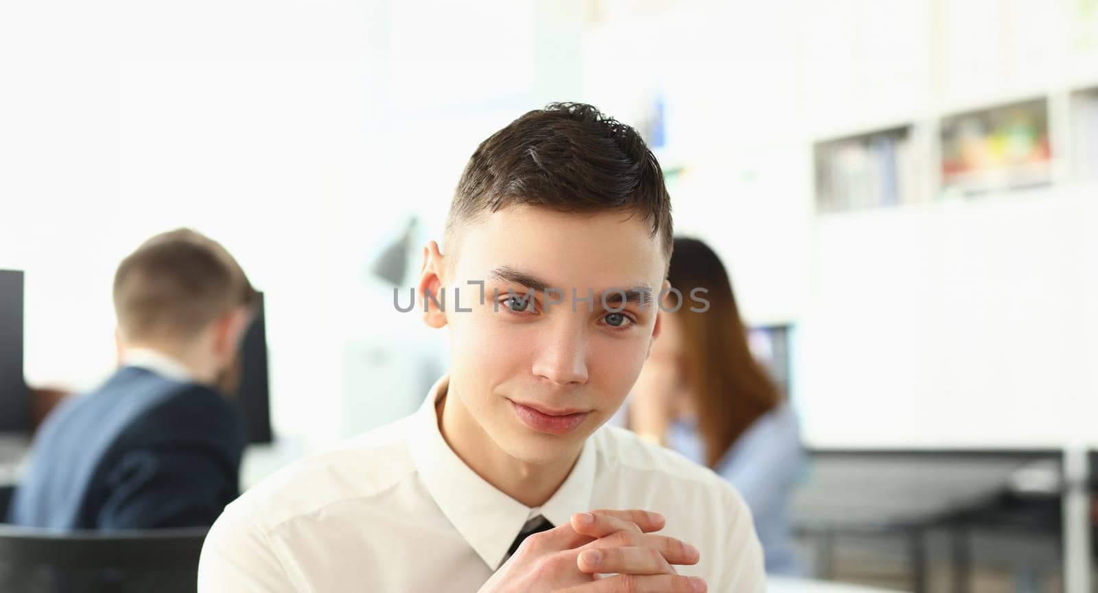 Portrait of unexperienced employee looking at camera with big joy. Man in classy tie sitting in big modern office with colleagues talking about important business project. Company meeting concept